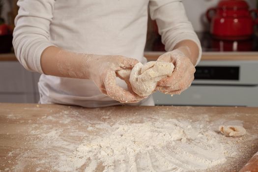 Charming little kid dressed in a white blouse is kneading the dough for baking a bread at a kitchen against a white wall with shelves and bulbs on it. Happy childhood concept.