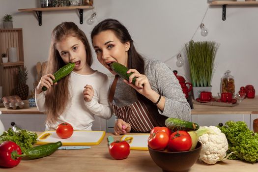 Happy loving family are cooking together. Good-looking mommy and her little girl are posing with tomato and cucumber and having fun at the kitchen, against a white wall with shelves and bulbs on it. Homemade food and little helper.