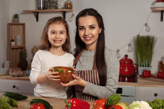 Happy loving family are cooking together. Smiling mom and her kid are making a vegetable salad and smiling at the kitchen, against a white wall with shelves and bulbs on it. Homemade food and little helper.