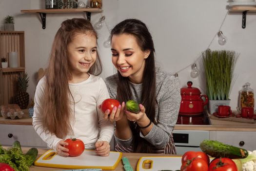 Happy loving family are cooking together. Delightful mom and her kid are posing with tomato and cucumber at the kitchen, against a white wall with shelves and bulbs on it. Homemade food and little helper.