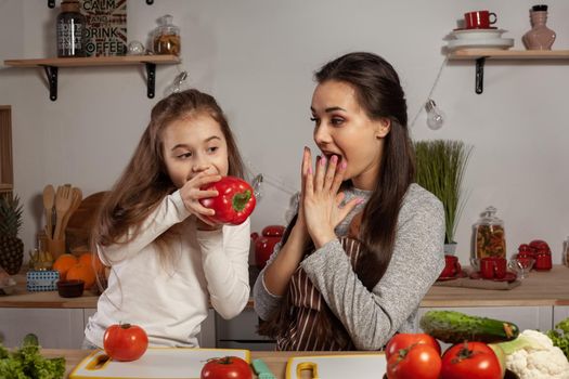 Happy loving family are cooking together. Loving mother and her little princess are making a vegetable salad and tasting a red pepper at the kitchen, against a white wall with shelves and bulbs on it. Homemade food and little helper.