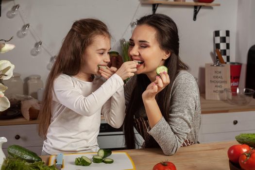 Happy loving family are cooking together. Attractive mother and her little princess are making a vegetable salad and tasting a cucumber at the kitchen, against a white wall with shelves and bulbs on it. Homemade food and little helper.