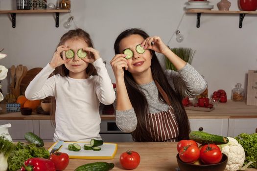 Happy loving family are cooking together. Alluring mum and her child are making a vegetable salad and fooling around at the kitchen, against a white wall with shelves and bulbs on it. Homemade food and little helper.