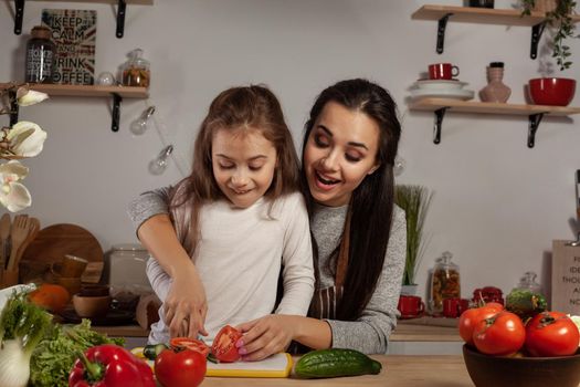 Happy loving family are cooking together. Pretty mum and her child are making a vegetable salad and having fun at the kitchen, against a white wall with shelves and bulbs on it. Homemade food and little helper.