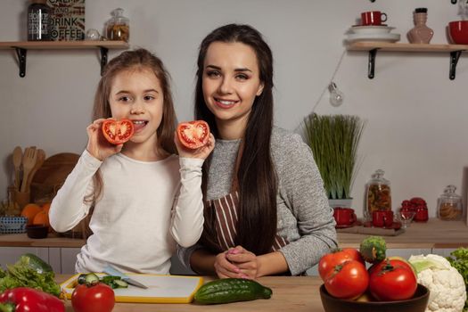 Happy loving family are cooking together. Pretty mother and her daughter are making a vegetable salad and posing with tomato at the kitchen, against a white wall with shelves and bulbs on it. Homemade food and little helper.