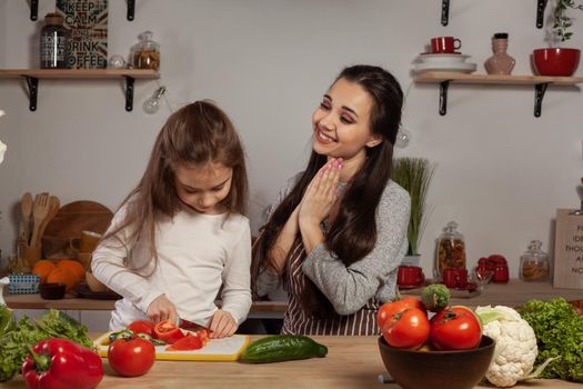 Happy loving family are cooking together. Gorgeous mom and her kid are making a vegetable salad and mommy is touched by her offspring at the kitchen, against a white wall with shelves and bulbs on it. Homemade food and little helper.