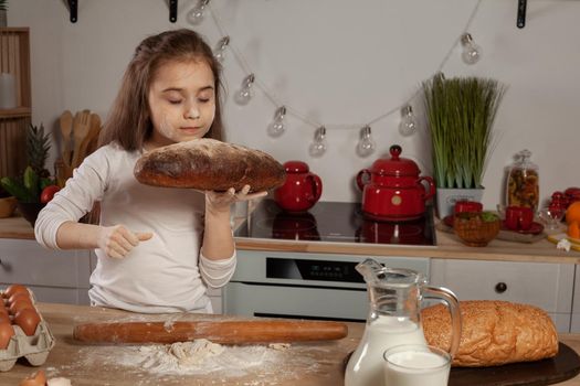 Beautiful little brunette kid dressed in a white blouse is siffing a bread at a kitchen against a white wall with shelves and bulbs on it. Happy childhood concept.