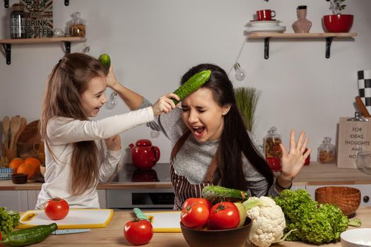 Happy loving family are cooking together. Wonderful mother and her daughter are fighting each other by cucumbers and having fun at the kitchen, against a white wall with shelves and bulbs on it. Homemade food and little helper.