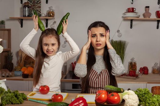 Happy loving family are cooking together. Brunette mother and her little princess are making a vegetable salad and tired of each other at the kitchen, against a white wall with shelves and bulbs on it. Homemade food and little helper.
