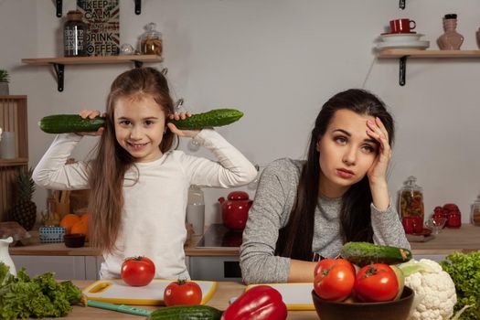 Happy loving family are cooking together. Brunette mommy and her little girl are making a vegetable salad and tired of each other at the kitchen, against a white wall with shelves and bulbs on it. Homemade food and little helper.