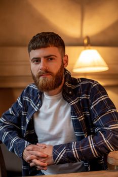 Portrait handsome bearded man wearing plaid shirt at modern cafe. Guy sitting in wooden chair and relaxing.