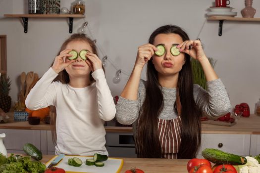 Happy loving family are cooking together. Alluring mommy and her little girl are making a vegetable salad and fooling around at the kitchen, against a white wall with shelves and bulbs on it. Homemade food and little helper.