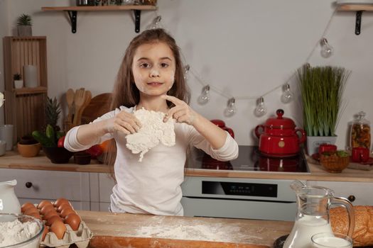 Pretty little brunette kid dressed in a white blouse is posing with a dough for baking bread and looking at the camera at kitchen against a white wall with shelves and bulbs on it. Happy childhood concept.