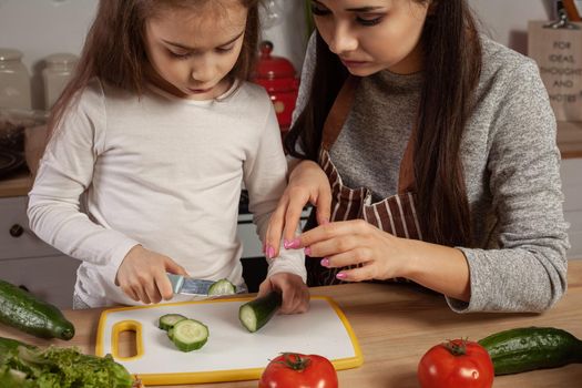 Happy loving family are cooking together. Attractive mom and her kid are making a vegetable salad at the kitchen, against a white wall with shelves and bulbs on it. Homemade food and little helper.