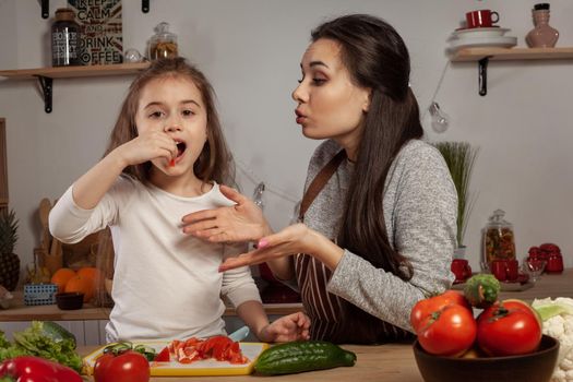 Happy loving family are cooking together. Beautiful mommy and her little girl are making a vegetable salad and tasting it at the kitchen, against a white wall with shelves and bulbs on it. Homemade food and little helper.