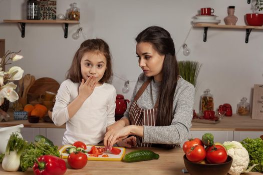 Happy loving family are cooking together. Charming mum and her child are making a vegetable salad and having fun at the kitchen, against a white wall with shelves and bulbs on it. Homemade food and little helper.