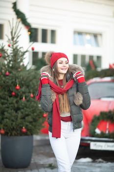 Pleasant fashionable winter girl smiling posing at red vintage car surrounded by snowflakes medium shot. Beautiful young woman enjoying december Christmas holiday having positive emotion