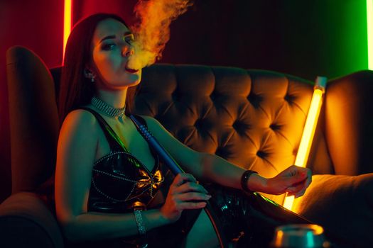 Lovely brunette female with a perfect make-up, in a sparkling necklace, leather dress and patent boots is smoking a hookah exhaling a smoke and looking at the camera sitting on an arm-chair at a luxury night club. Concept of pleasant spending time. Dark background with colorful neon backlights.