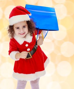 Laughing a little, curly-haired girl in a coat and hat of Santa Claus carries on his shoulder a colorful shopping bags. Close-up.Winter brown abstract background with white snowflakes.