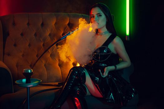 Sexy brunette lady with a perfect make-up, in a sparkling necklace, leather dress and patent boots is smoking a hookah exhaling a smoke sitting on an arm-chair and looking at the camera at a luxury night club. Concept of pleasant spending time. Dark background with colorful neon backlights.