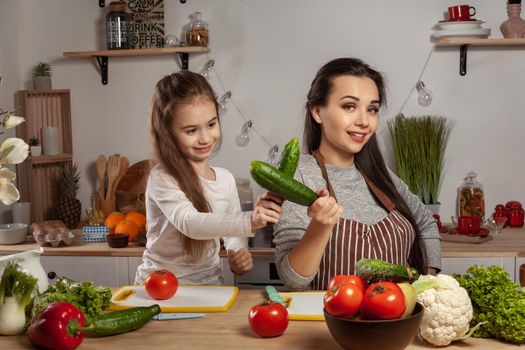 Happy loving family are cooking together. Good-looking mother and her little princess are posing with tomato and cucumber and having fun at the kitchen, against a white wall with shelves and bulbs on it. Homemade food and little helper.