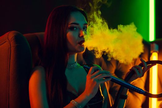 Close-up shot of a wonderful brunette female with a perfect make-up, in a sparkling necklace and a leather dress smoking a hookah exhaling a smoke sitting sideways on an arm-chair at a luxury night club. Concept of pleasant spending time. Dark background with colorful neon backlights.