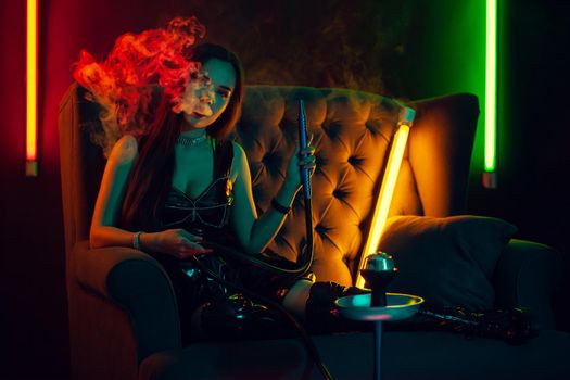 Amazing brunette female with a perfect make-up, in a sparkling necklace, leather dress and patent boots is smoking a hookah exhaling a smoke and looking at the camera sitting on an arm-chair at a luxury night club. Concept of pleasant spending time. Dark background with colorful neon backlights.