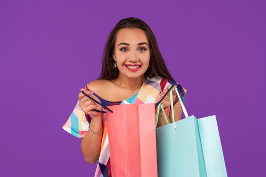 Beautiful emotional woman peeks into a package, in the hands of multi-colored shopping bags on a purple background. Studio shot. Copy space