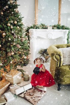 Stock photo portrait of adorable girl in red dress with festive print holding beautifully wrapped golden present in hands while sitting on the floor next to decorated Christmas treee with garland..