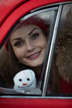 Pleasant fashionable winter girl smiling posing at red vintage car surrounded by snowflakes medium shot. Beautiful young woman enjoying december Christmas holiday having positive emotion