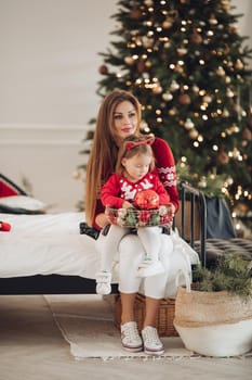 Stock photo of loving mother in green dress giving her little daughter in pyjama dress a Christmas present. They are next to beautifully decorated Christmas tree under snowfall.