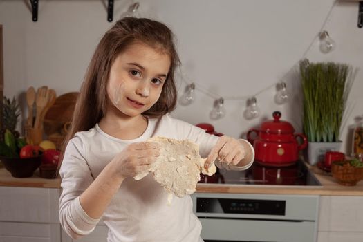 Pretty little brunette girl dressed in a white blouse is posing with a dough for baking a bread at a kitchen against a white wall with shelves and bulbs on it. Happy childhood concept.