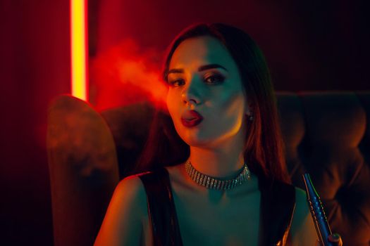 Close-up shot of a dazzling brunette lady with a perfect make-up, in a sparkling necklace and a leather dress smoking a hookah exhaling a smoke sitting on an arm-chair at a luxury night club. Concept of pleasant spending time. Dark background with colorful neon backlights.