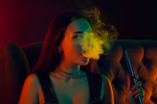 Close-up shot of a dazzling brunette girl with a perfect make-up, in a sparkling necklace, leather dress and patent boots smoking a hookah exhaling a smoke sitting on an arm-chair at a luxury night club. Concept of pleasant spending time. Dark background with colorful neon backlights.