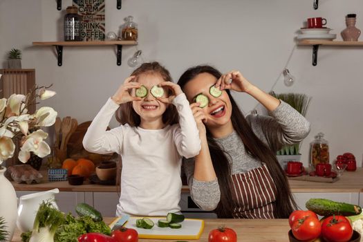Happy loving family are cooking together. Alluring mom and her kid are making a vegetable salad and fooling around at the kitchen, against a white wall with shelves and bulbs on it. Homemade food and little helper.