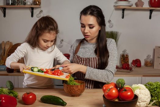 Happy loving family are cooking together. Beautiful mum and her child are making a vegetable salad and having fun at the kitchen, against a white wall with shelves and bulbs on it. Homemade food and little helper.