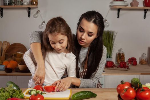 Happy loving family are cooking together. Gorgeous mommy and her little girl are making a vegetable salad and having fun at the kitchen, against a white wall with shelves and bulbs on it. Homemade food and little helper.