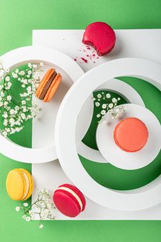 Beautiful festive composition macaron or macaroon cookies with white circles and flowers on green background from above, colorful almond small cakes, modern greetings card, top view