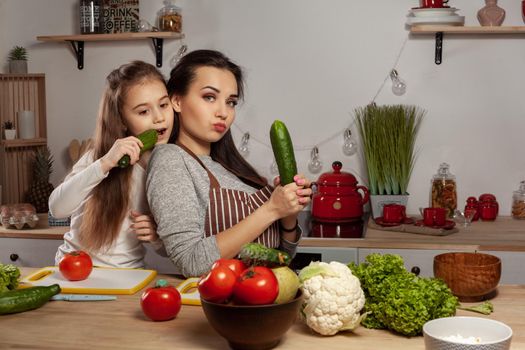 Happy loving family are cooking together. Good-looking mum and her child are posing with tomato and cucumber and looking at the camera at the kitchen, against a white wall with shelves and bulbs on it. Homemade food and little helper.