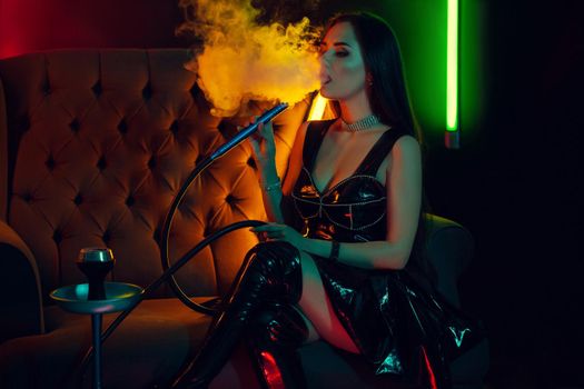 Charming brunette girl with a perfect make-up, in a sparkling necklace, leather dress and patent boots is smoking a hookah exhaling a smoke sitting sideways on a sofa at a luxury night club. Concept of pleasant spending time. Dark background with colorful neon backlights.