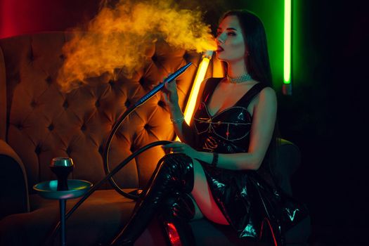 Charming brunette model with a perfect make-up, in a sparkling necklace, leather dress and patent boots is smoking a hookah exhaling a smoke sitting sideways on an arm-chair at a luxury night club. Concept of pleasant spending time. Dark background with colorful neon backlights.