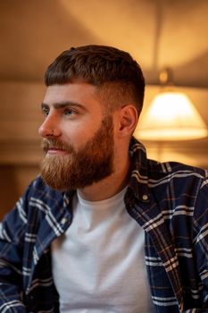 Portrait handsome bearded man wearing plaid shirt at modern cafe. Guy sitting in wooden chair and relaxing.