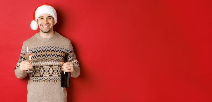 Concept of winter holidays, new year and celebration. Portrait of handsome man in santa hat and sweater, holding champagne and making toast on christmas party, standing over red background.