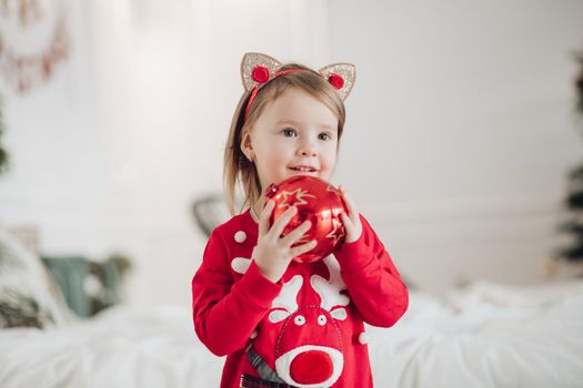 Stock photo portrait of adorable girl in red dress with festive print holding beautifully wrapped golden present in hands while sitting on the floor next to decorated Christmas treee with garland...