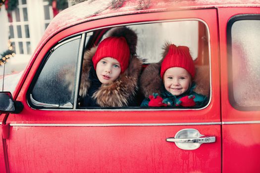 Smiling cute winter boy in red hat sitting in car having fun medium shot. Happy beautiful female baby in warm clothing having positive emotion outdoor surrounded by snowflakes enjoying childhood