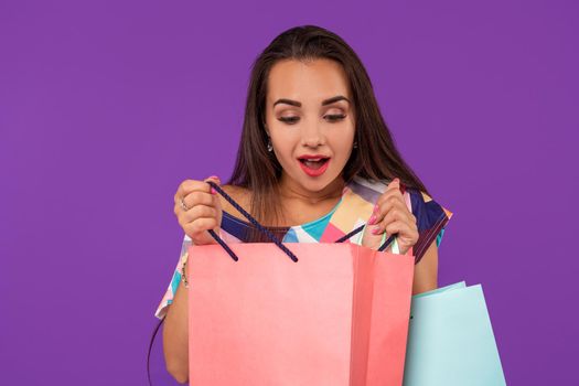 Beautiful emotional woman peeks into a package, in the hands of multi-colored shopping bags on a purple background. Studio shot. Copy space