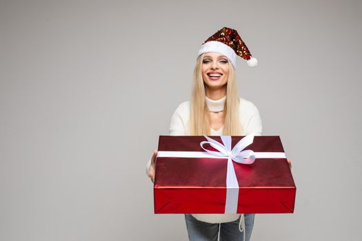 Stock photo of smiling cheerful blonde woman in Santa hat offering beautiful red wrapped Christmas gift with white bow to the camera. Isolated on white.
