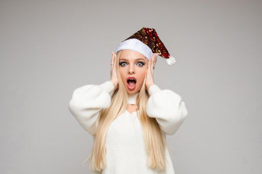 Stock photo of excited blonde girl in sparkling Santa hat in white sweater holding hands at face looking at camera with open mouth. Shocked or surprised concept. Isolated.