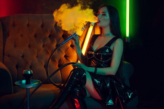 Charming brunette female with a perfect make-up, in a sparkling necklace, leather dress and patent boots is smoking a hookah exhaling a smoke sitting on an arm-chair at a luxury night club. Concept of pleasant spending time. Dark background with colorful neon backlights.