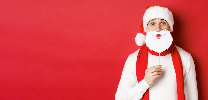 Concept of christmas, winter holidays and celebration. Portrait of funny man in santa hat, holding beard mask, enjoying new year party, standing over red background.